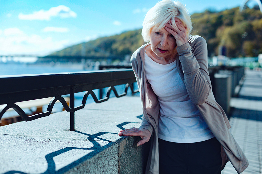 Need to take my pills. Exhausted older lady leaning on a barrier and touching her forehead while suffering from a terrible headache outdoors.
