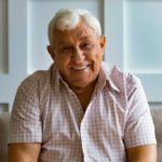 Close up headshot portrait of smiling mature man sit on couch at home look at camera posing for picture, happy positive senior male or optimistic grandfather feel good relax on comfortable sofa