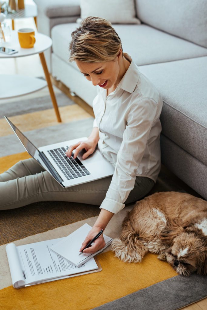 Smiling businesswoman working on laptop and taking notes while working at home. Her dog is sleeping next to her.