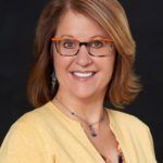 Woman with reddish-brown hair and glasses, smiling, and wearing a yellow sweater.