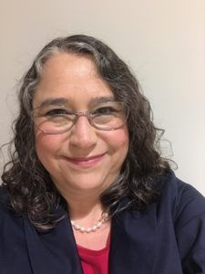Woman with dark brown hair and glasses, smiling, and wearing a navy blue jacket and red shirt.